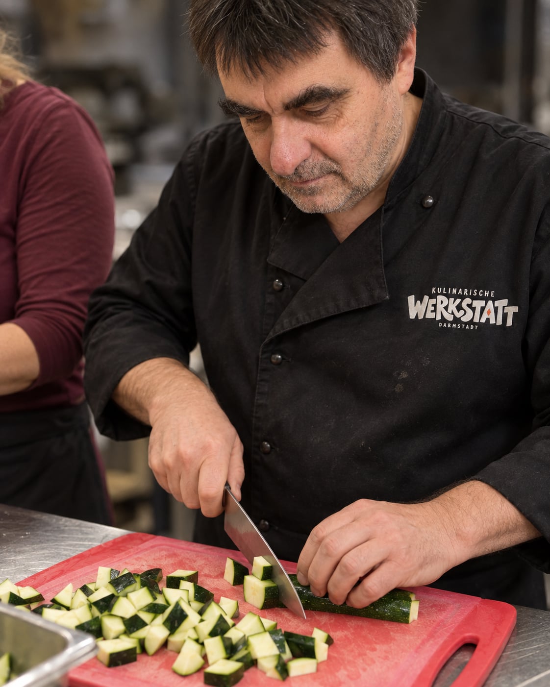 Alf schneidet Zucchini auf einem roten Brett, daneben arbeitet ein Teilnehmer