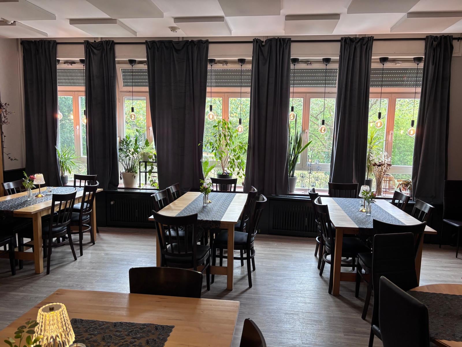 Originalaufnahme der Fensterseite mit Holztischen, dunklen Stühlen und Vorhängen